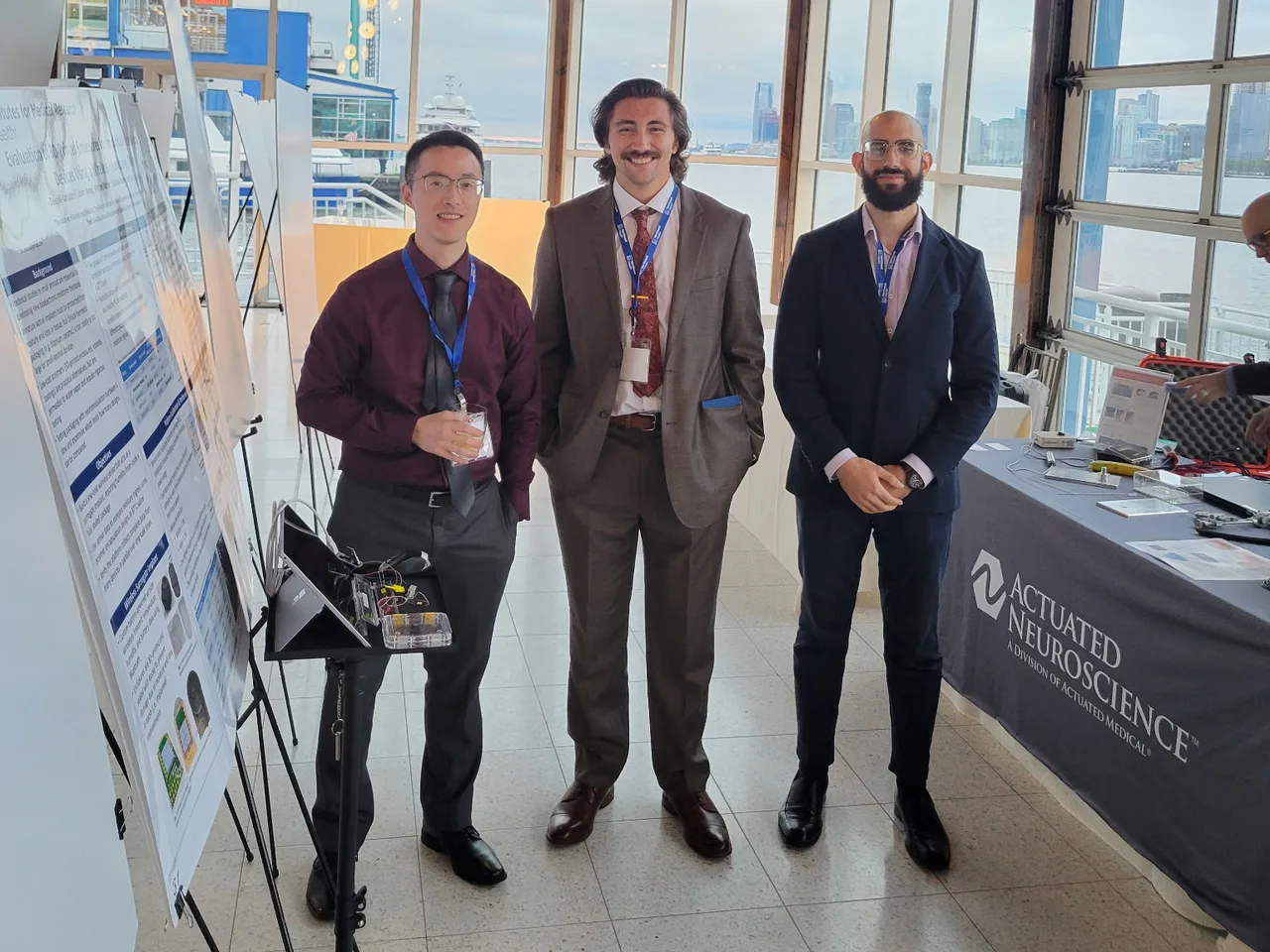 Three lab members standing beside a research poster at the summit, with the Hudson River through the windows behind them